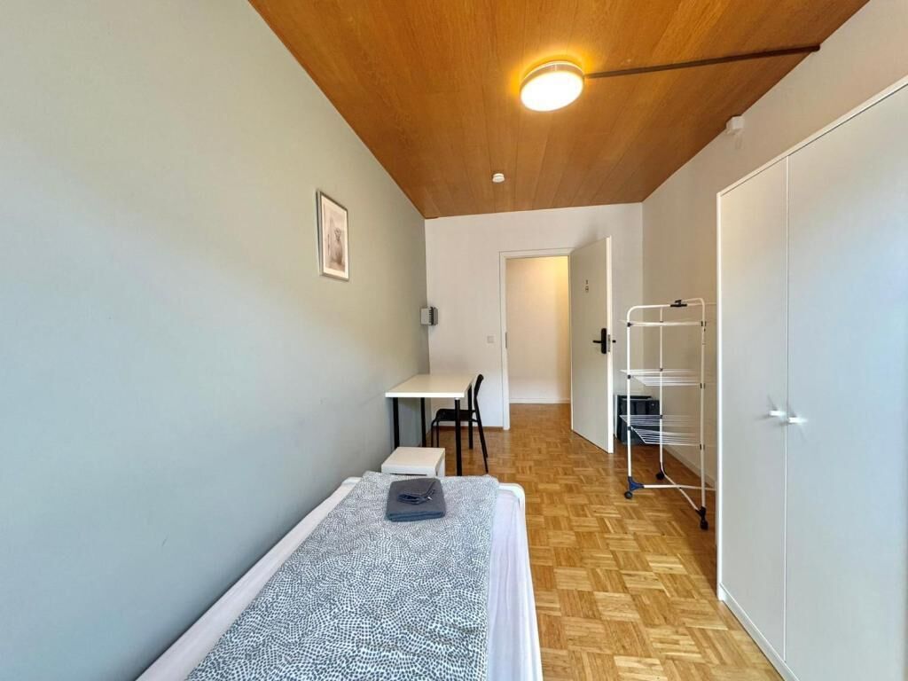 Una habitación pequeña y funcional con cama, escritorio y silla. Se observa una puerta que conduce a otra habitación y una pizarra en la pared.