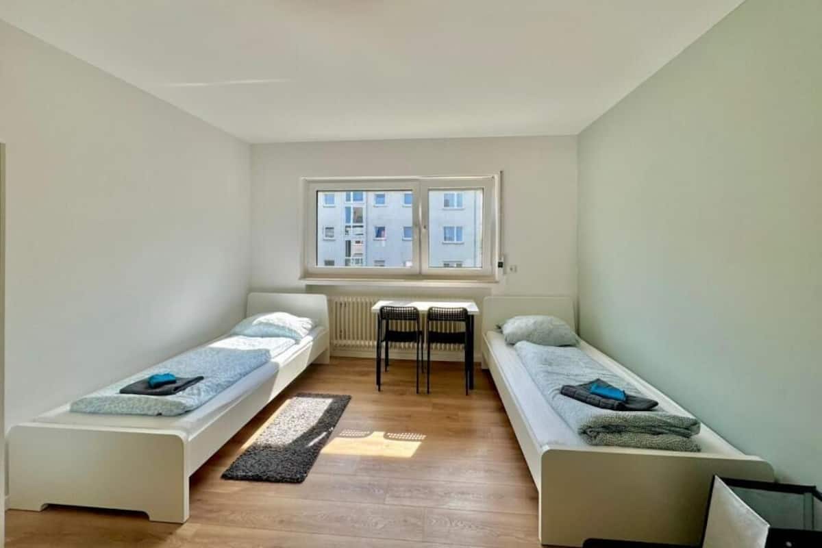 A compact room with two single beds, a small table, and a window providing a view of buildings outside.