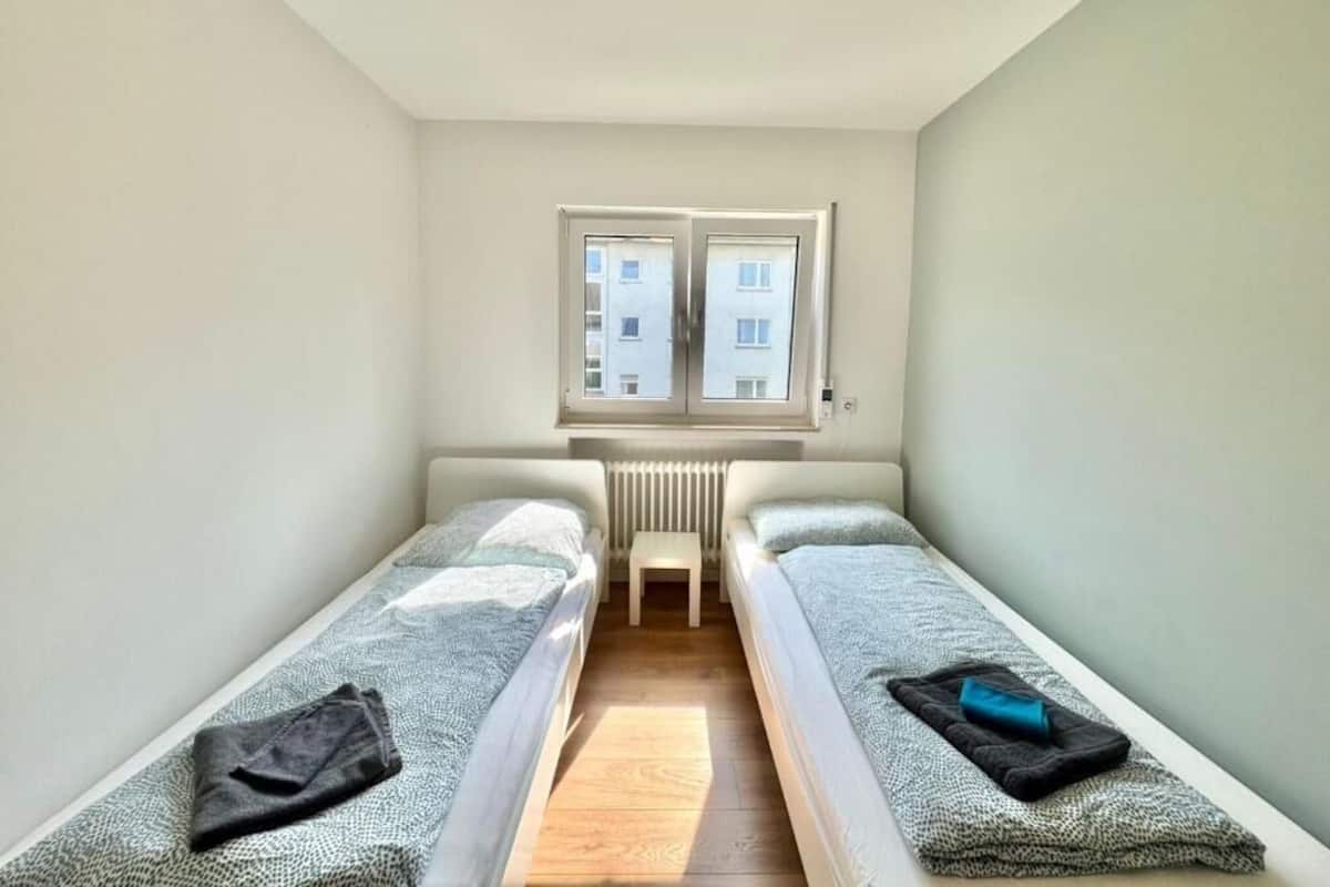 A small, narrow room with two single beds, a radiator, and a window with a view of buildings.