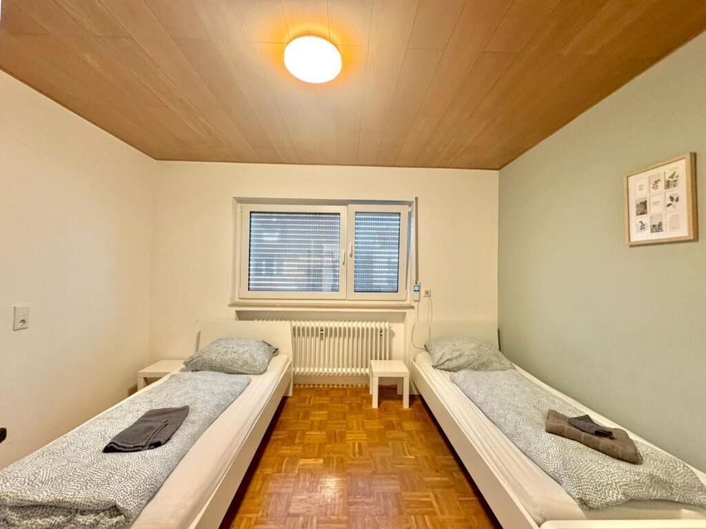 A small, cozy room with two single beds, a radiator, a window with blinds, and a ceiling light.