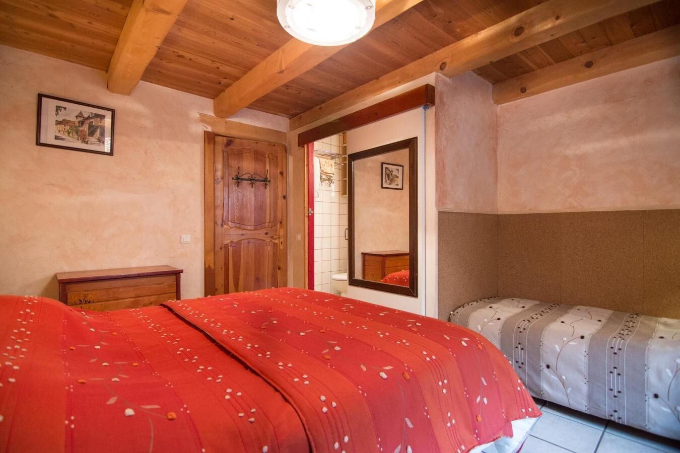 A bedroom with a wooden ceiling, a bed with a red bedspread, a wooden dresser, and a mirror on the wall.