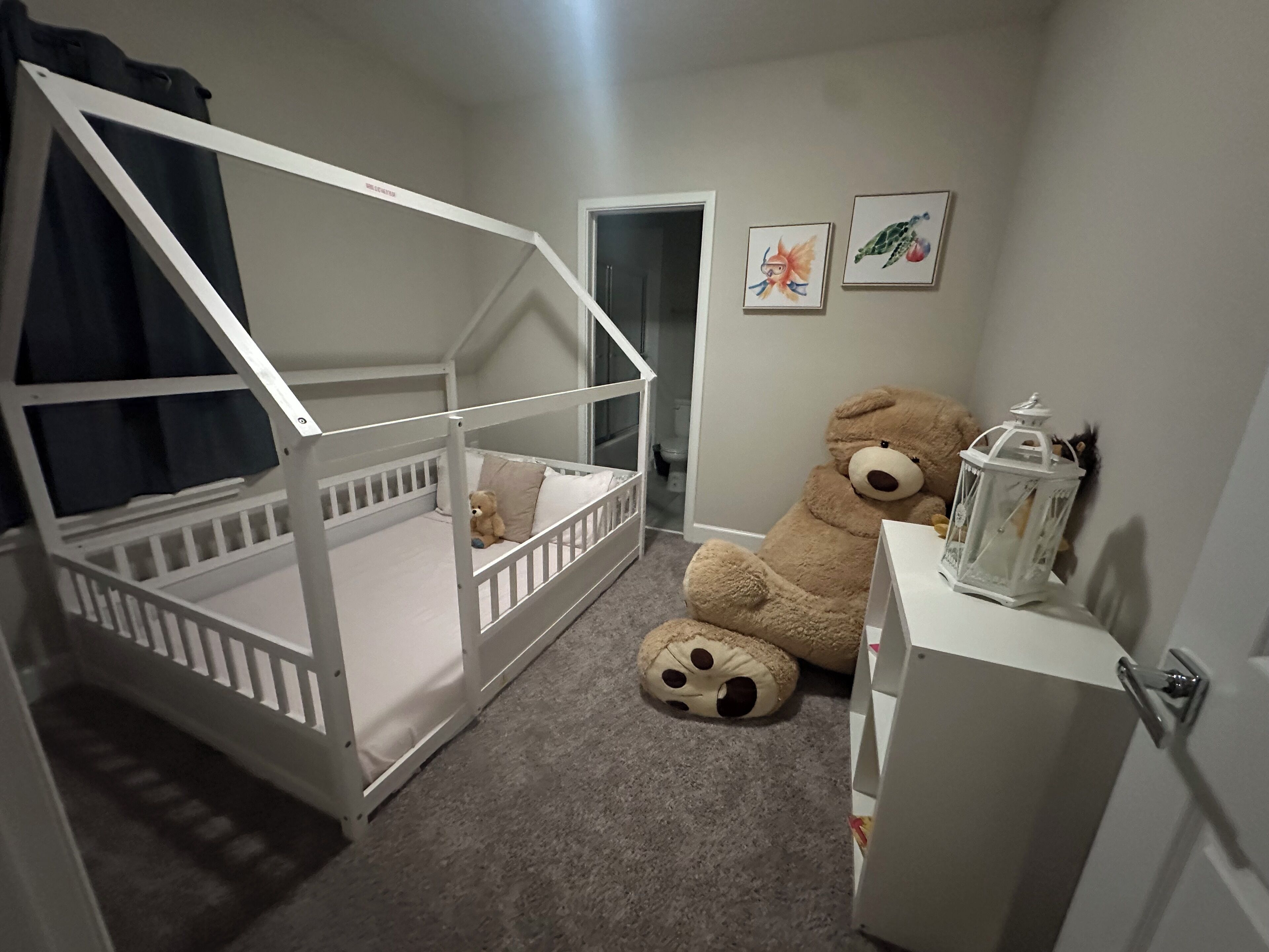 Ein Kinderzimmer mit einem Etagenbett, einem großen Teddybären, einem kleinen Tisch mit einer Laterne und zwei gerahmten Bildern an der Wand.