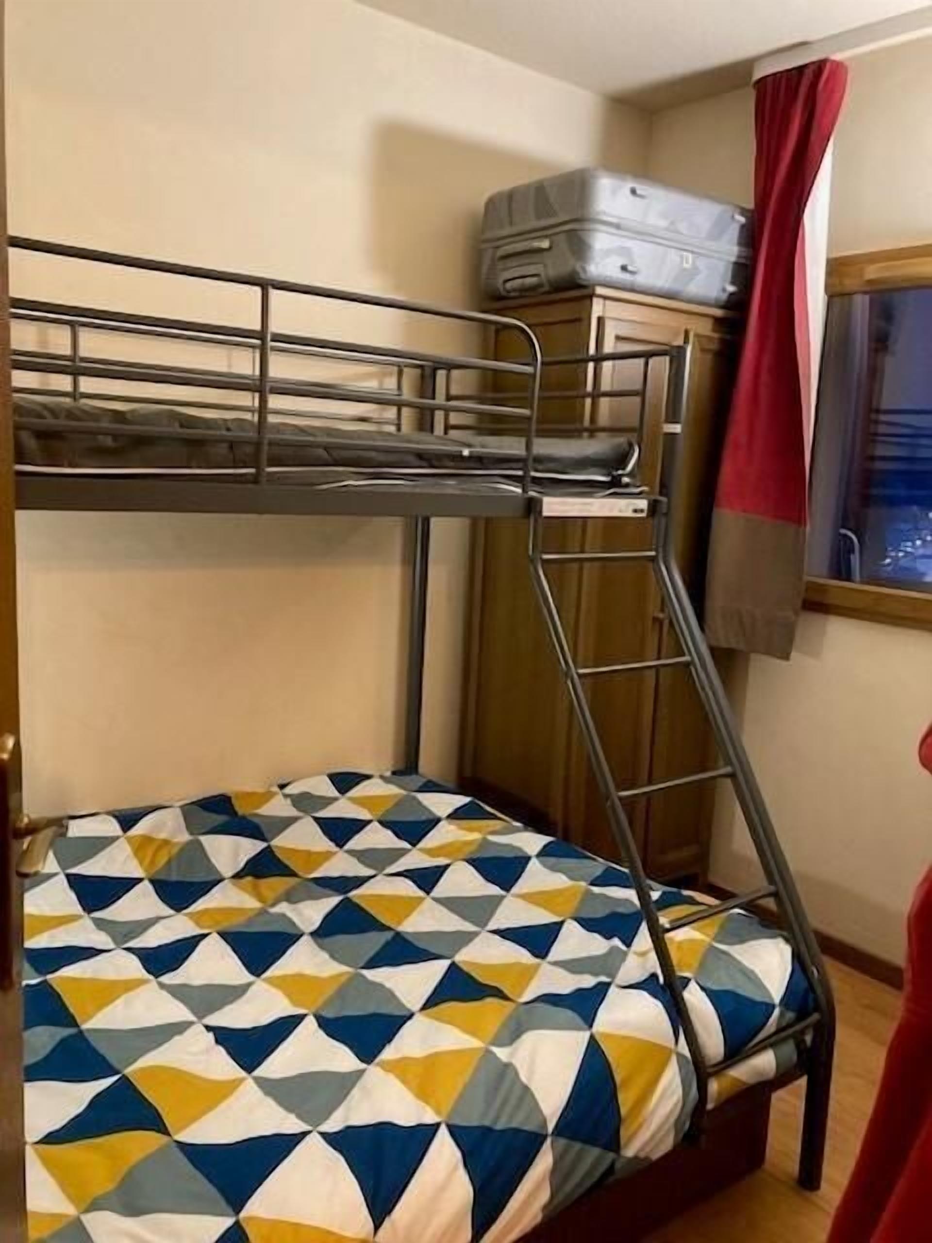 Ein Stockbett mit einem zart gemusterten Bettbezug, ein Koffer auf dem oberen Bett und ein Fenster mit roten Vorhängen.