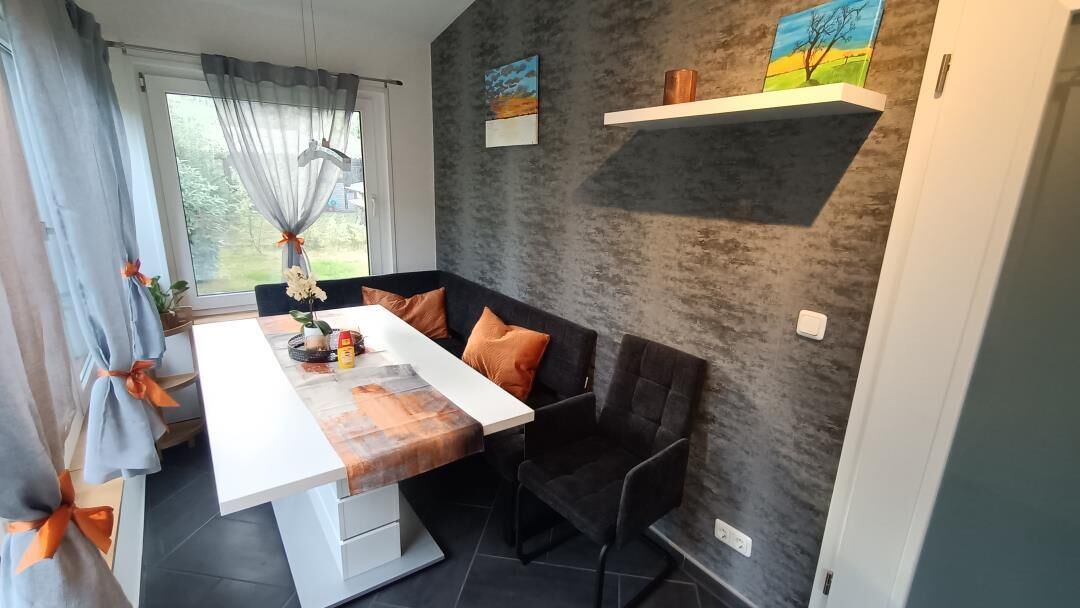 A dining area with a white table, black chairs, and orange cushions. There are two paintings on the wall and a shelf with a vase.