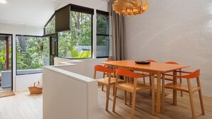 A modern dining area with a wooden table and orange chairs, large windows with a view of greenery, and a hanging light fixture.