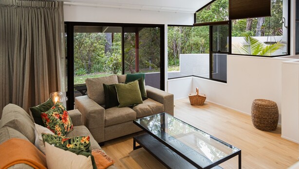 A modern living room with a sofa, a glass coffee table, and a view of a lush garden through large windows.