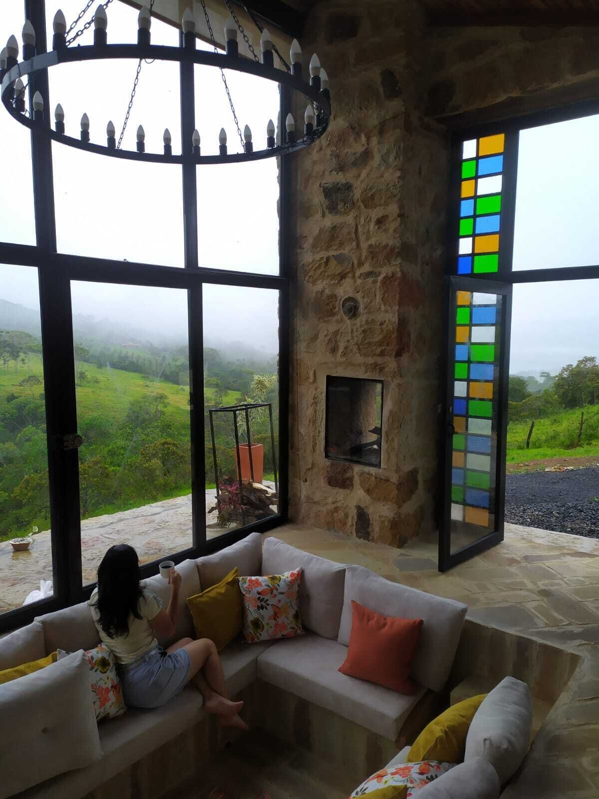 Une pièce chaleureuse avec un mur en pierre, une cheminée et de grandes fenêtres donnant sur un paysage verdoyant.