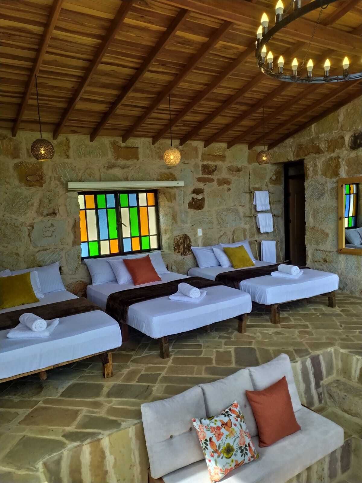 A room with stone walls, wooden ceiling beams, and a large window with colorful panes. There are three beds with white linens and multiple pillows. A sofa with a floral cushion is in the foreground.