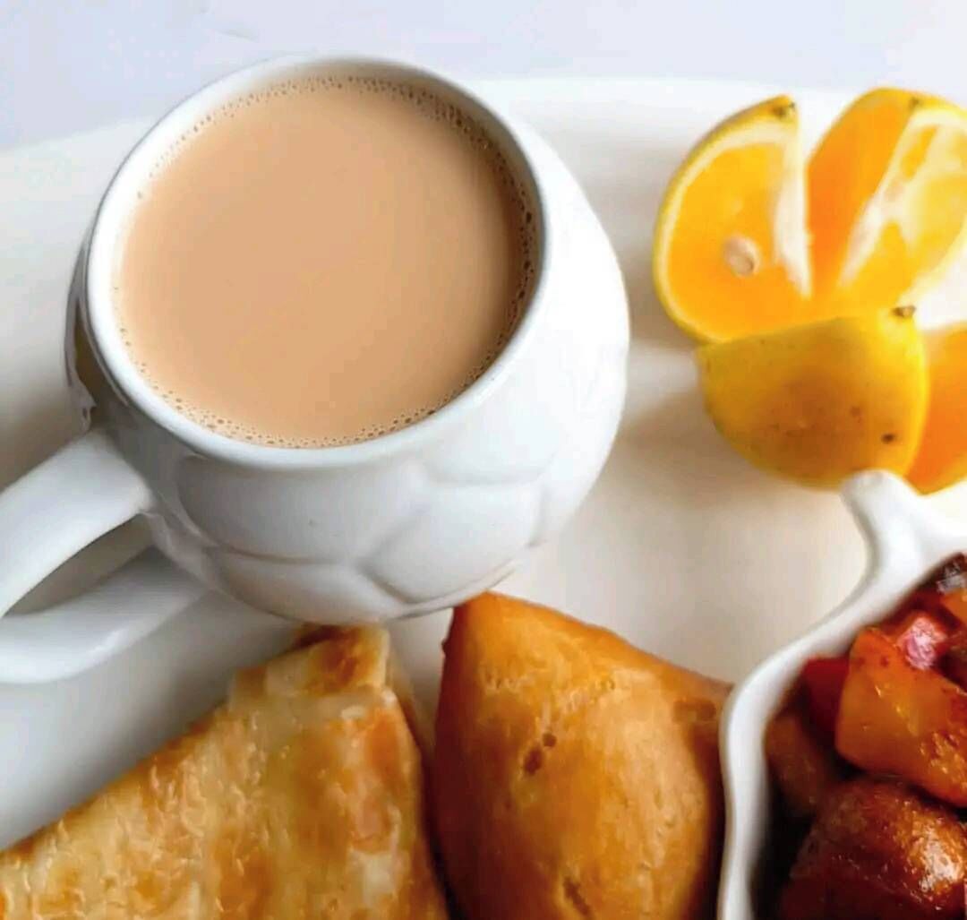 Una taza blanca llena de una bebida marrón claro, junto a un plato de comida que incluye un pastel horneado y papas asadas.