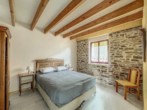 A bedroom with a wooden ceiling, a stone wall, a bed with a headboard, a nightstand, a chair, and a window with curtains.