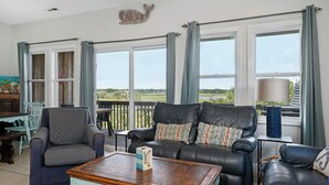 A living room with a view of the countryside, featuring a black leather sofa, a grey armchair, and a wooden coffee table.