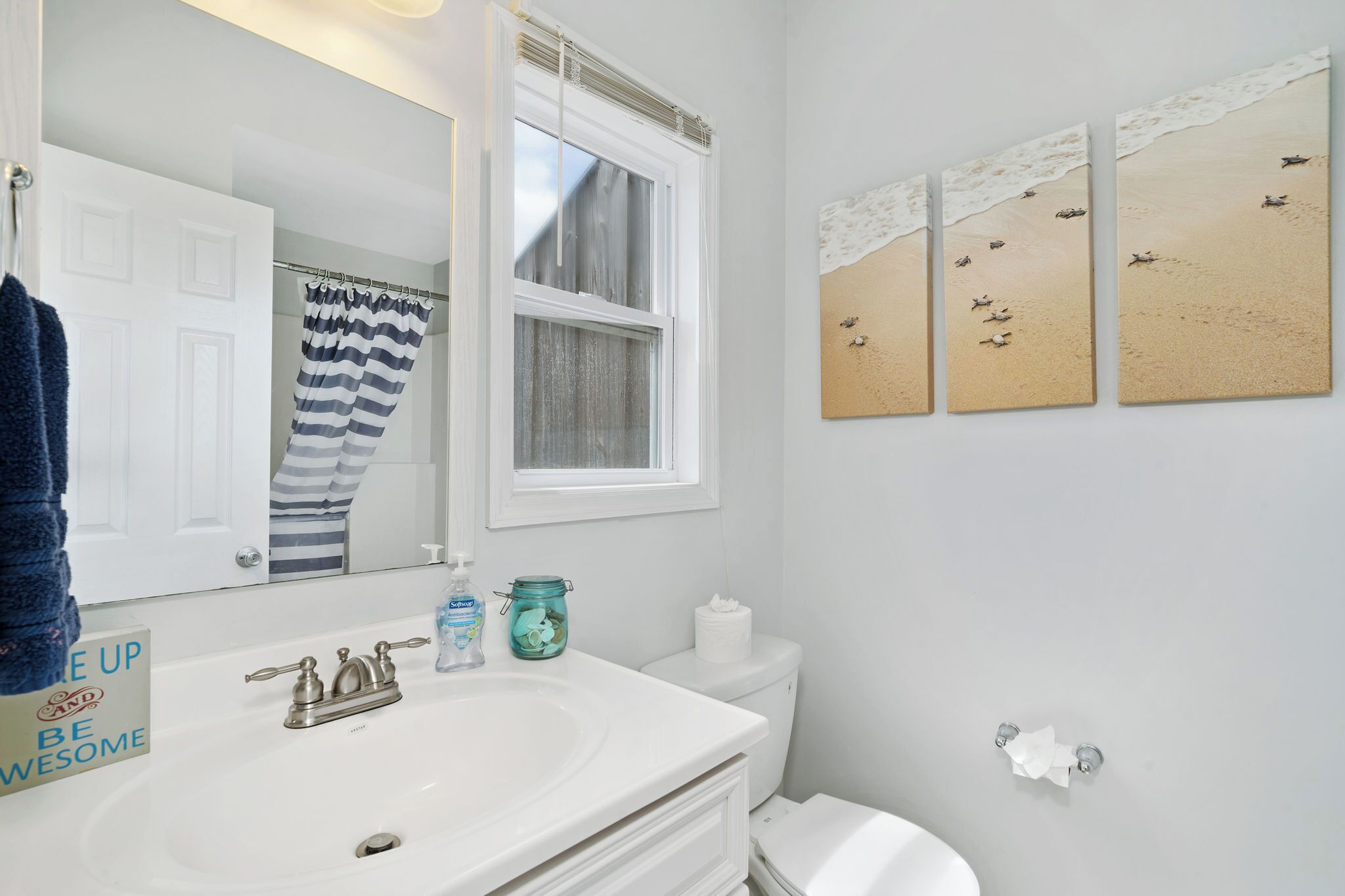 A bathroom with a white sink, a mirror, a striped shower curtain, and three beach-themed wall art pieces.