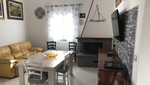 A dining area with a white table and chairs, a yellow sofa, a fireplace, and a TV mounted on the wall.