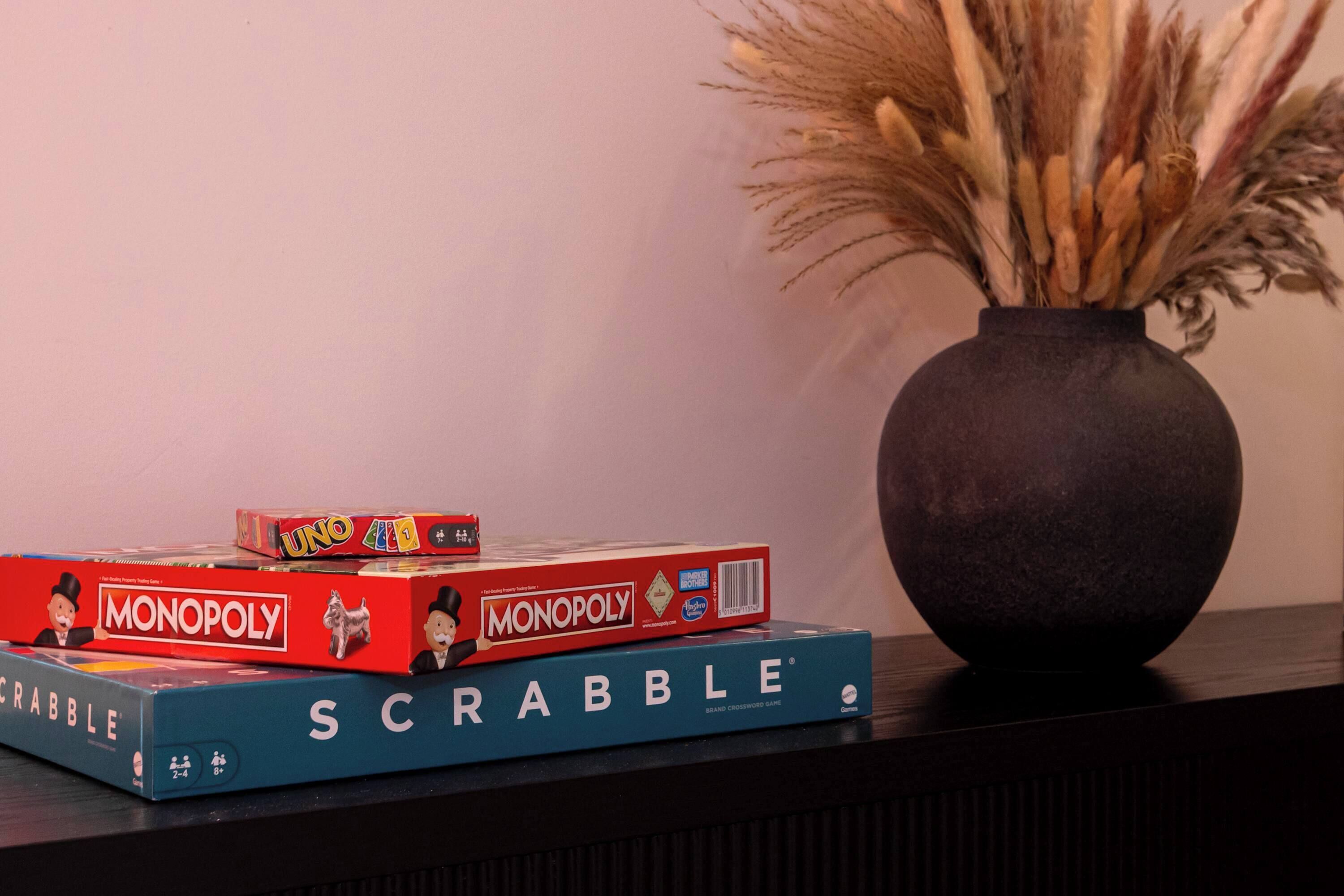A stack of board games including Monopoly and Scrabble, with a vase of dried plants beside them.