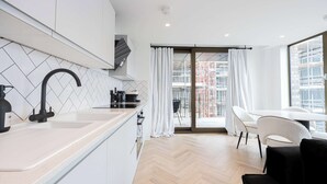 Een moderne keuken met een visgraatvloer, een groot raam met witte gordijnen en een eethoek met een witte tafel en stoelen.