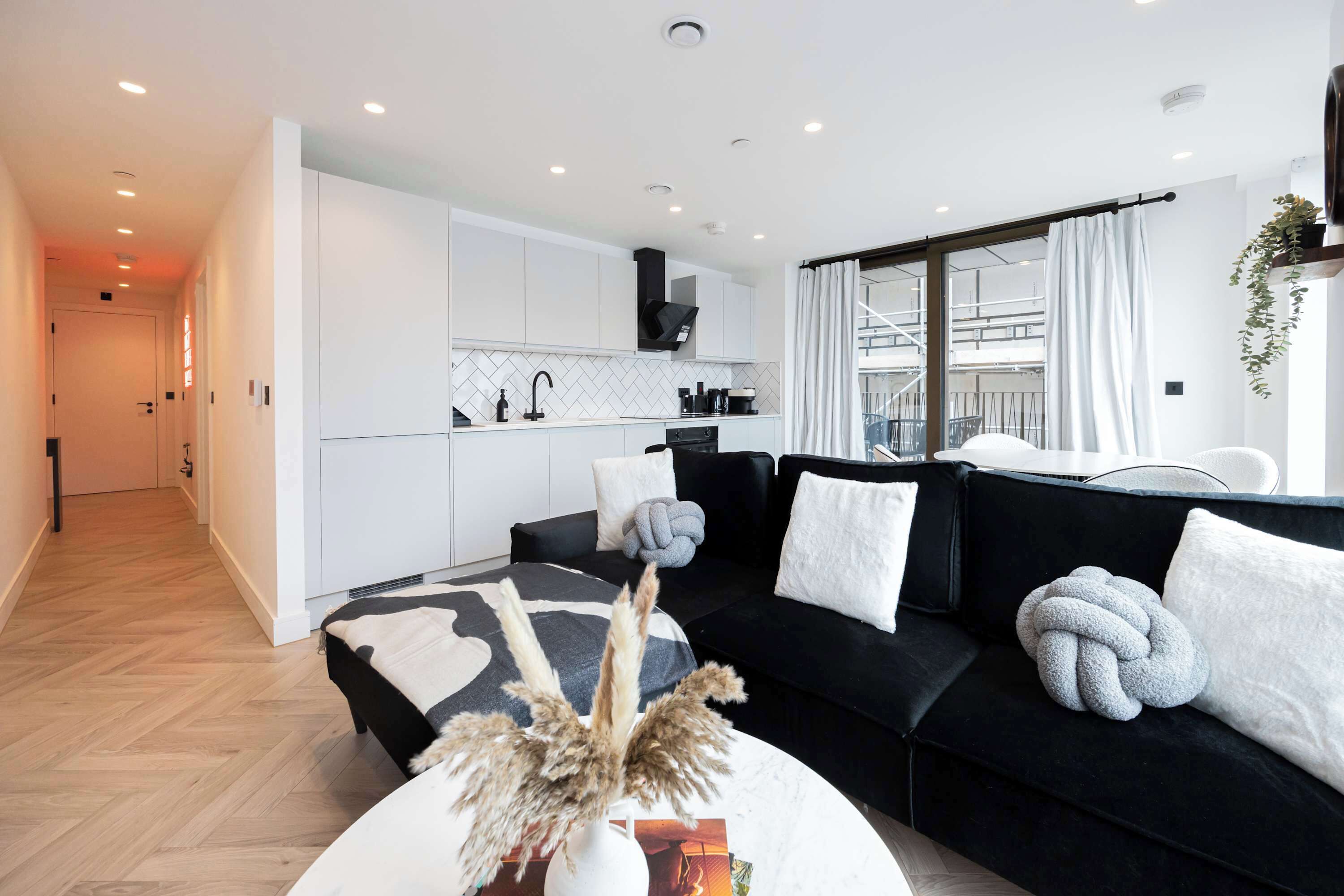 A modern living room with a black sofa, white cushions, and a coffee table with dried plants. A kitchen with white cabinets and a black range hood is visible in the background.