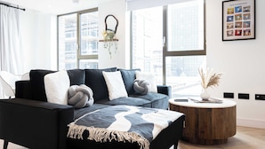 Een moderne woonkamer met een zwarte hoekbank, een ronde houten salontafel en grote ramen met uitzicht op de stad.