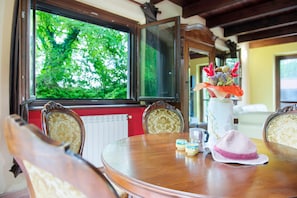 Un coin repas avec une table ronde, des chaises, un vase de fleurs, un chapeau et une fenêtre donnant sur des arbres.