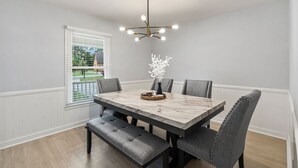 Une salle à manger avec une table en marbre, six chaises grises à revêtement rembourré, un banc et un lustre moderne.