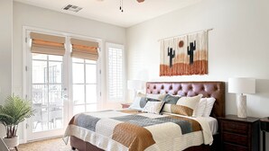 A bedroom with a large bed, a nightstand, a lamp, a potted plant, and a view of the outside. - Stylish Desert King Room w/ Balcony + Pool View (La Quinta)