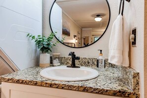 Une salle de bain avec un comptoir en granit, un lavabo blanc, un miroir rond, une plante, un distributeur de savon et une serviette accrochée au mur.