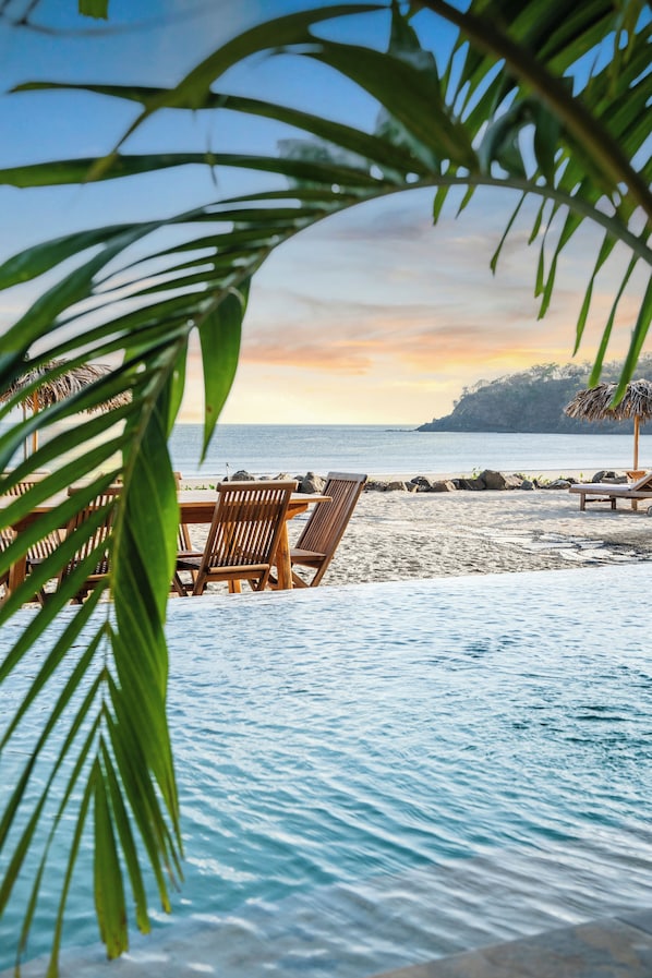 A beachside setting with a wooden table and chairs, a pool, and a view of the ocean and a distant mountain.