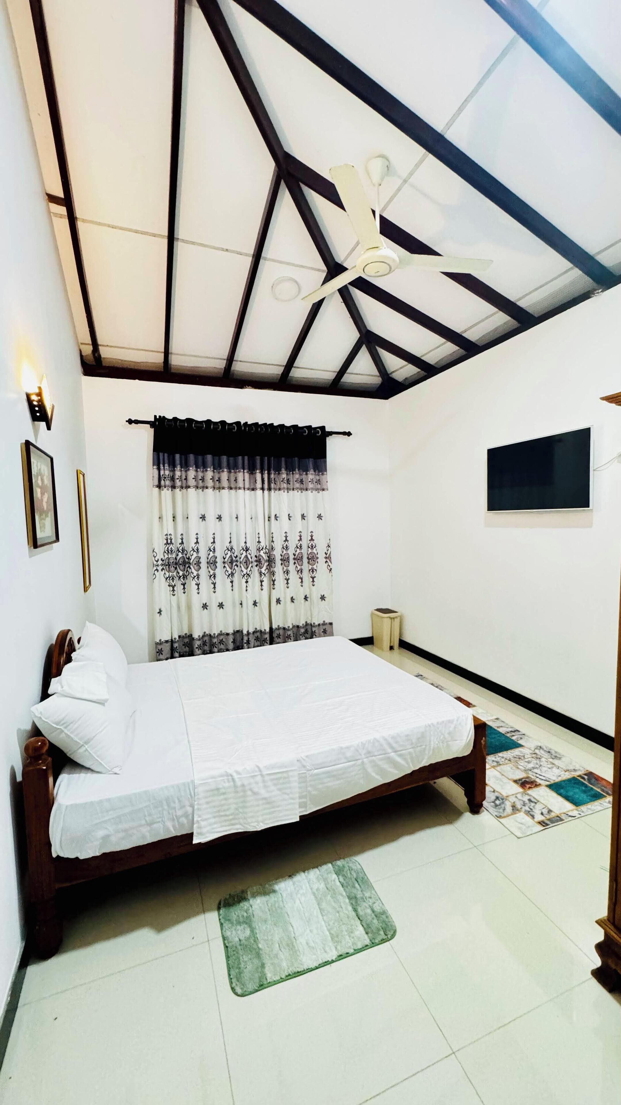 A bedroom with a bed, a ceiling fan, a window with curtains, a bedside table, and a wall-mounted TV.