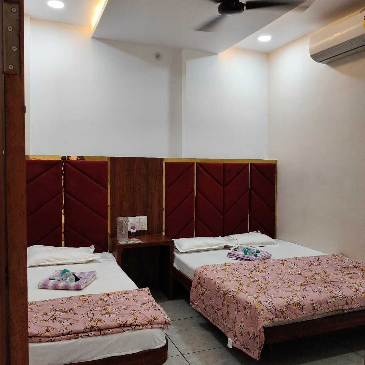 Habitación de hotel con dos camas, cada una con cabecera de madera y colcha estampada. Hay una mesita de noche entre las camas, y un equipo de aire acondicionado montado en la pared.