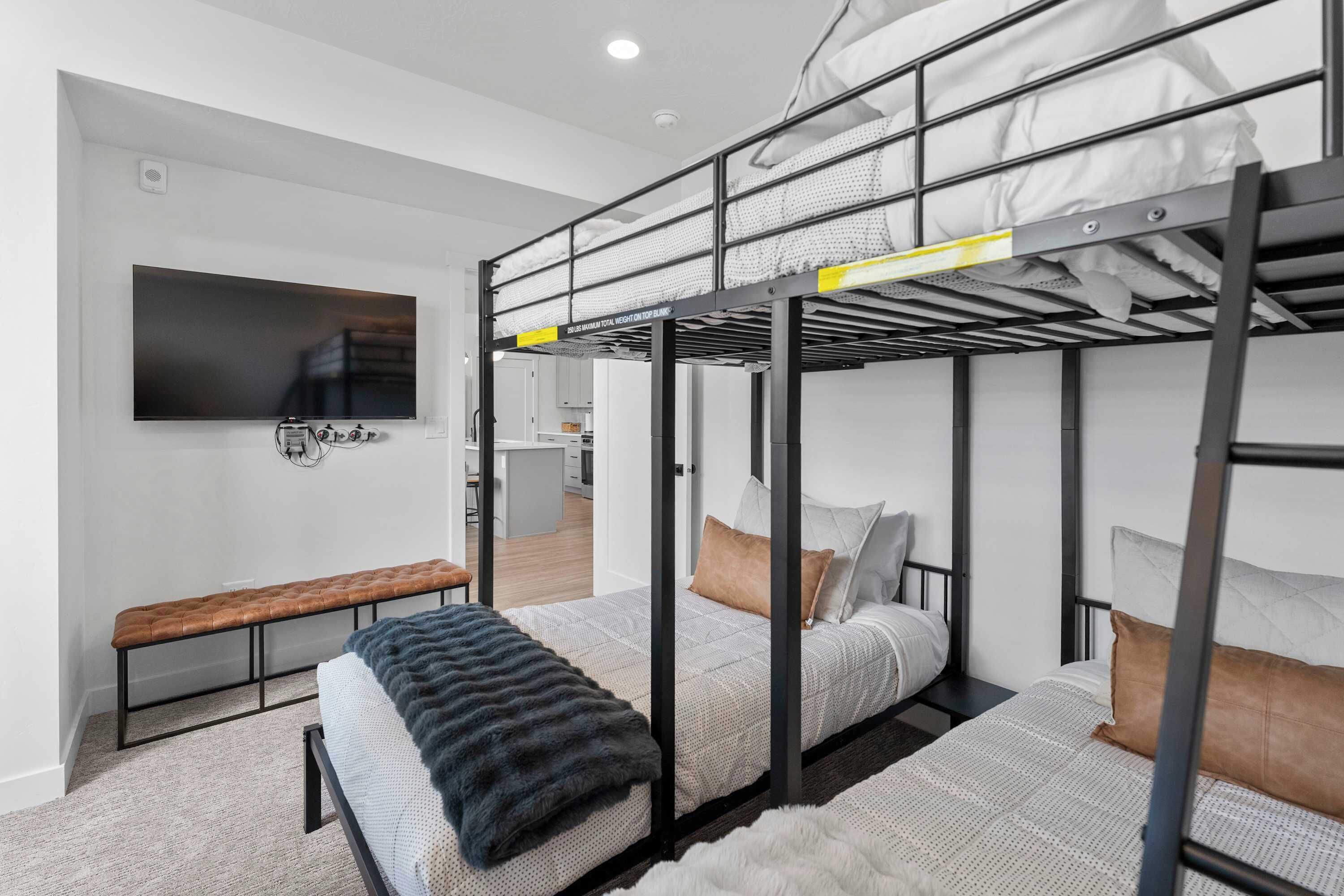 A modern, compact room with bunk beds, a flat-screen TV, a bench, and a kitchenette in the background.