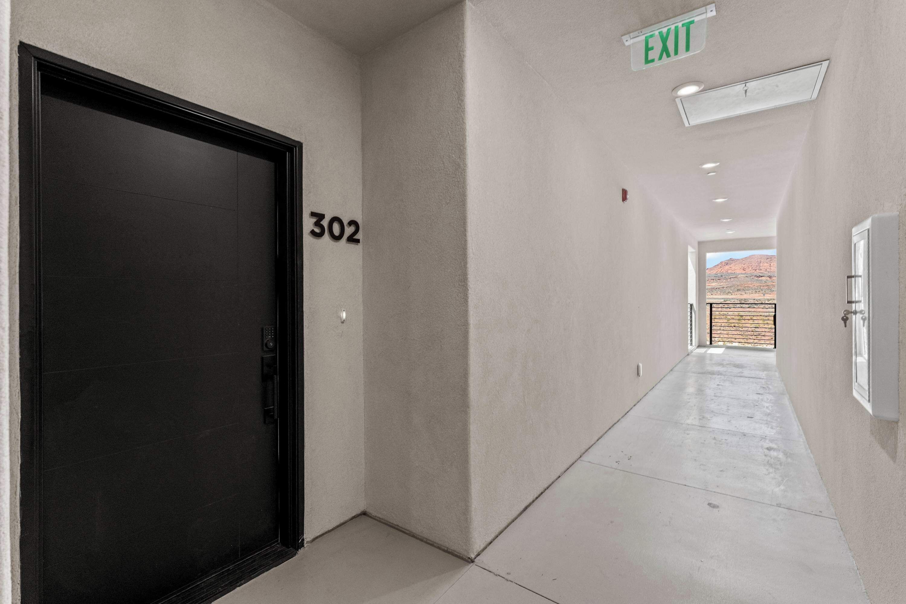 Un pasillo con una puerta negra marcada '302', un letrero de salida y una vista de paisaje desértico a través de una puerta abierta al final.