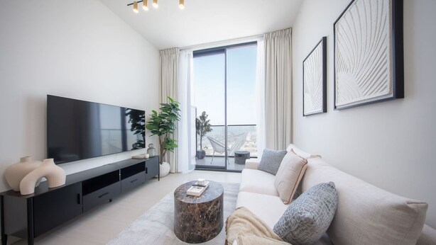 Un salon moderne doté d’un grand téléviseur à écran plat, d’un meuble de rangement noir, d’une table basse ronde, d’un canapé beige et d’une grande fenêtre avec des rideaux.