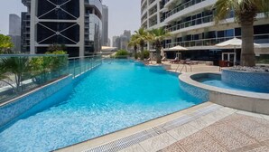 A swimming pool with a tiled edge, surrounded by palm trees and lounge chairs, with high-rise buildings in the background. - Vibrant Studio in JLT Sleeps 4 With Lake Views (Dubai)