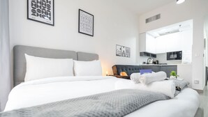 A modern hotel room with a large bed, a bedside table with a lamp, a small kitchen area, and artwork on the walls. - Cozy JVC Studio for 2 With Balcony (Dubai)