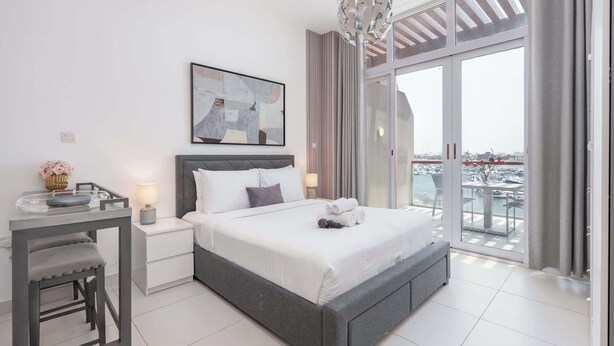 A modern bedroom with a large bed, a desk, and a view of the marina through the balcony doors. - Chic Studio in Palm Jumeirah Sleeps 2 With Balcony (Dubai)