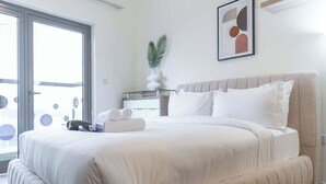 A modern hotel room with a large bed, a bedside table with a lamp, a framed abstract art piece, and a view through the glass door. - JVC Apartment That Sleeps 4 With a Great Pool (Dubai)