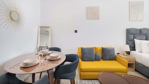 A modern living room with a round dining table, two blue chairs, a yellow sofa with grey cushions, and a grey armchair. - JVC Studio for 3 People With Swimming Pool (Dubai)