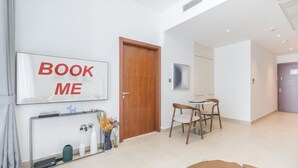 Ein modernes Wohnzimmer mit einem großen Fernseher, der "BOOK ME" anzeigt, einer Holztür, einem Essbereich mit Tisch und Stühlen sowie einem Konsolentisch mit Dekoration.