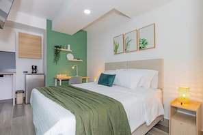 Una habitación de hotel moderna con cama, mesita de noche, escritorio y una kitchenette pequeña.