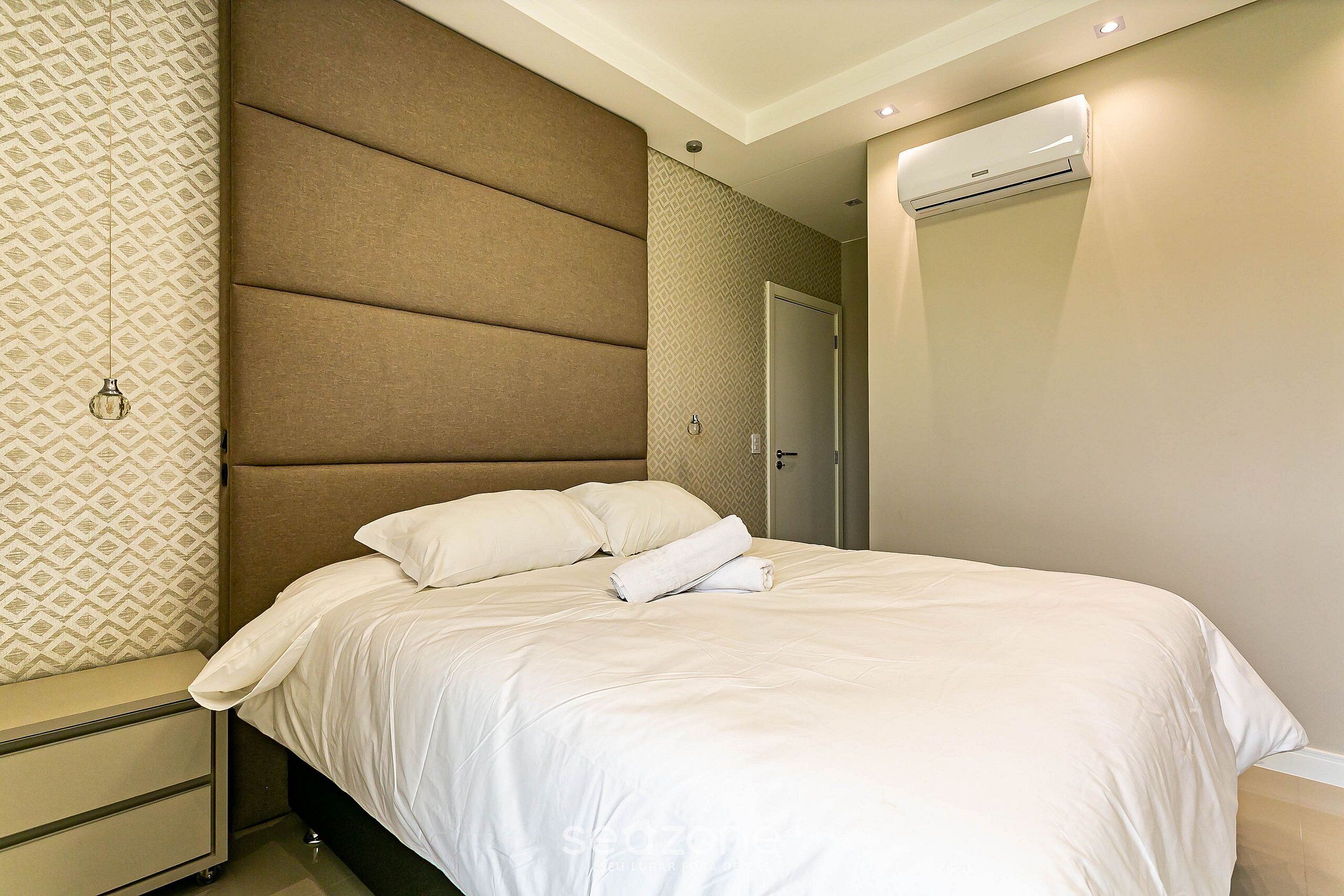 A neatly made bed with white linens and a folded towel, a bedside table with a drawer, and a wall-mounted air conditioning unit.