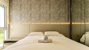 A neatly made bed with a rolled towel, a bedside table, and a window with a view of greenery. - Sophisticated fit With 3 Suites Jurere Les403 (Florianópolis)
