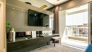 A modern living room with a large flat-screen TV mounted on the wall, a built-in console table with a vase and remote controls, a small stool, and a view of the city through large windows. - Sophisticated fit With 3 Suites Jurere Les403 (Florianópolis)