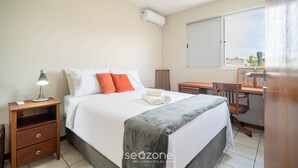 A hotel room with a bed, a desk, a chair, a nightstand, and an air conditioning unit. - Apt w/ Balcony & BBQ 450m From the sea Reh0203 (Florianópolis)