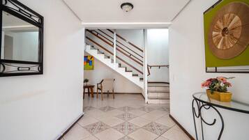 Un pasillo con suelo de baldosas, una escalera, un espejo, un adorno en la pared y una pequeña mesa con una planta en maceta.