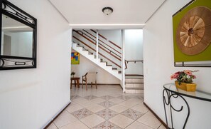 A hallway with a tiled floor, a staircase, a mirror, a wall-mounted decorative piece, and a small table with a potted plant. - Apt w/ Balcony & BBQ 450m From the sea Reh0203 (Florianópolis)