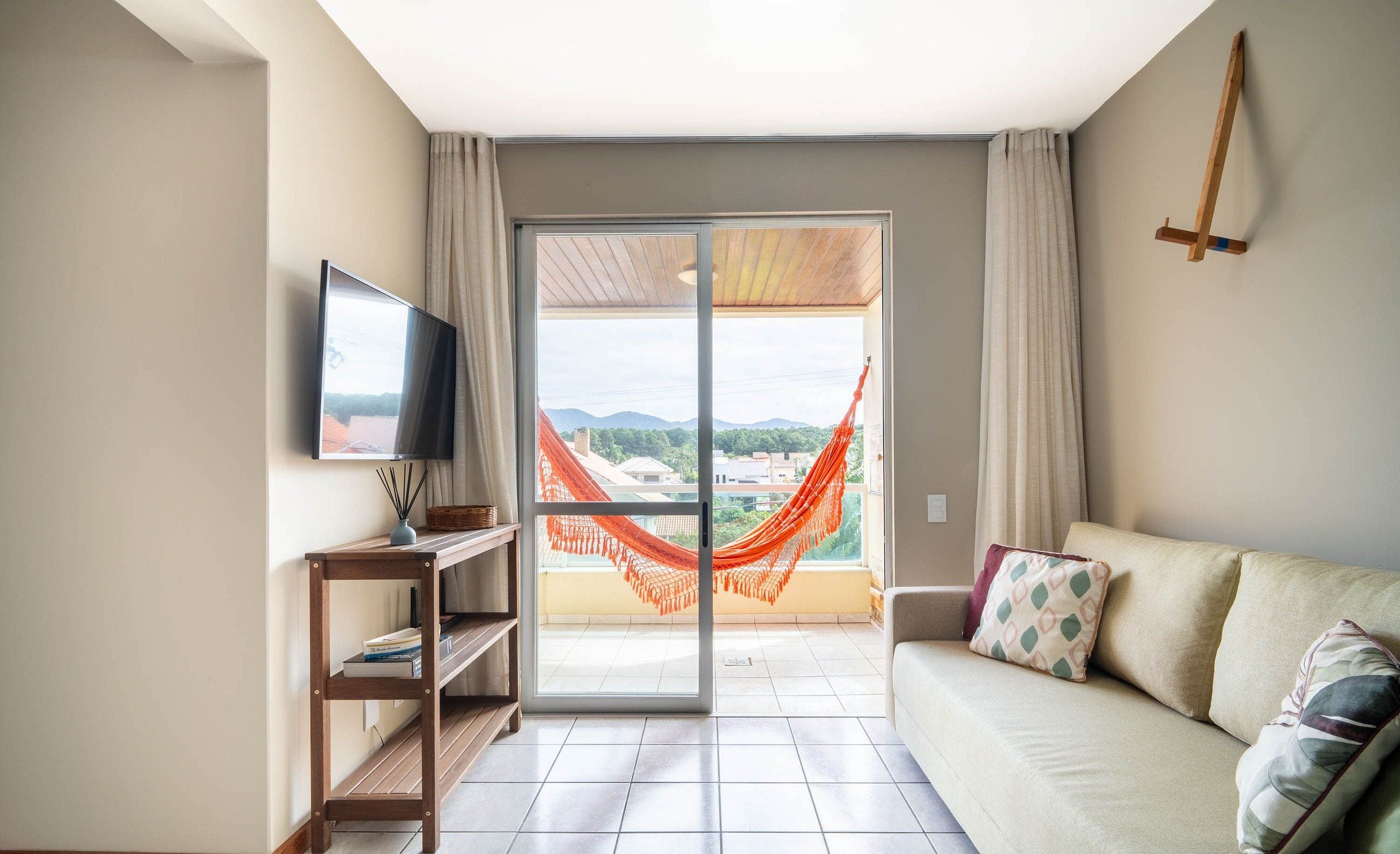 Sala de estar com sofá, televisão, prateleira de madeira, rede e varanda com vista.
