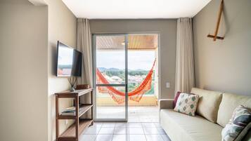 Sala de estar com sofá, televisão, prateleira de madeira, rede e varanda com vista.