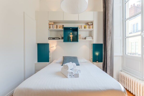 A neatly made bed with rolled towels, a shelf with books and a decorative item, and a window with curtains.
