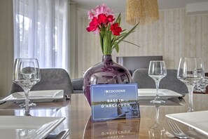 A dining table set with white plates, cutlery, and wine glasses, with a vase of pink flowers and a sign indicating 'Bassin d'Arcacchon'.