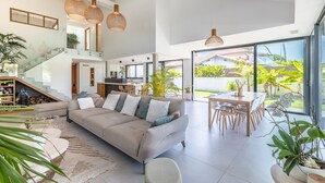 A spacious living area with a sofa, dining table, and chairs. There is a staircase with a glass railing leading to an upper level.