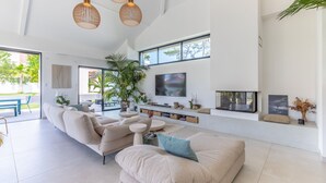 A spacious living room with a large sectional sofa, a coffee table, and a TV mounted on the wall. There are large windows providing natural light and a view of the outdoors.